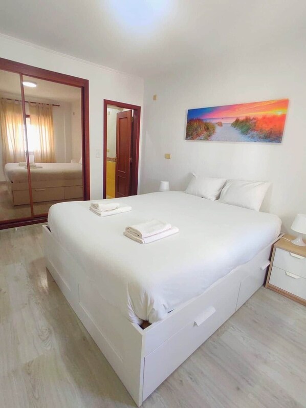 A hotel room with a large bed, a bedside table, a nightstand, a painting on the wall, and a door leading to another room. - The Golden Hour Retreat (Alcochete)