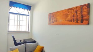 Un salon avec un canapé gris, deux coussins jaunes, un tapis marron, une fenêtre avec des bordures bleues et un tableau représentant des arbres.