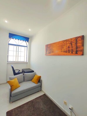 Ein Wohnzimmer mit einem grauen Sofa, zwei gelben Kissen, einem braunen Teppich, einem Fenster mit blauer Umrandung und einem Baumgemälde.