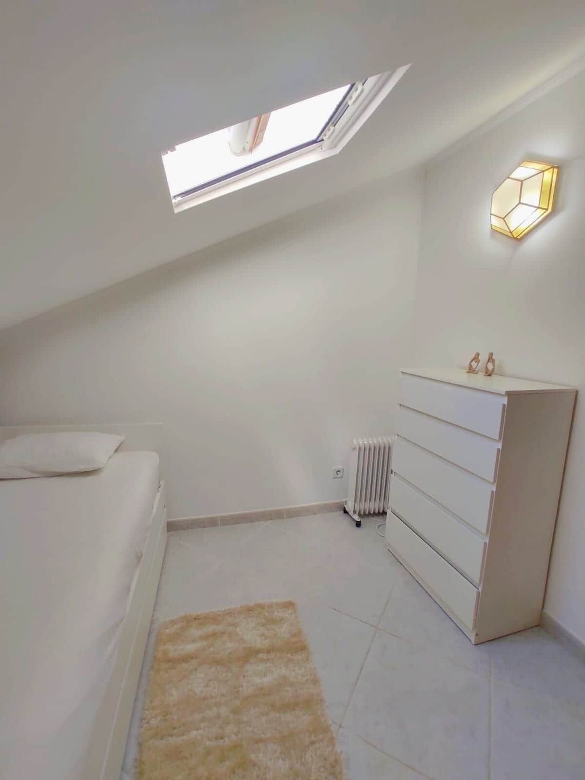 A bedroom with a skylight, a radiator, a white dresser, and a small rug.