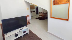 Un salon équipé d’un téléviseur à écran plat, d’un meuble TV en bois, d’un coin repas avec une table et des chaises, et d’une cuisine en arrière-plan.