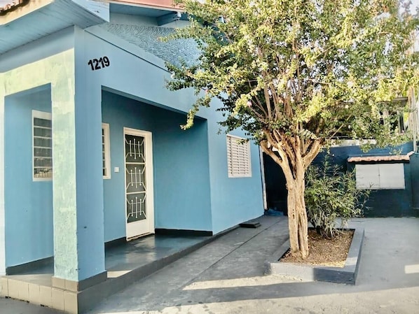 A light blue building with a number '1219' on the door, a tree in front, and a small bush to the right.