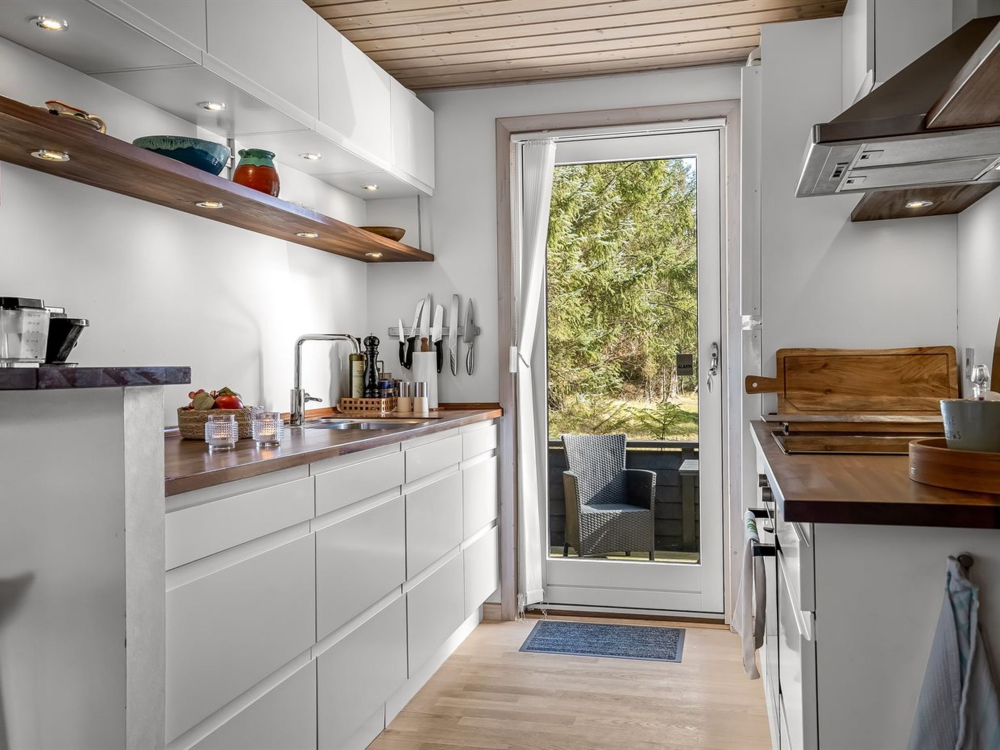 A modern kitchen with white cabinets, wooden shelves, and a view to the outside.