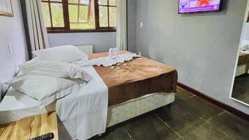 Un dormitorio con cama, televisor montado en la pared, ventana con cortinas y un escritorio de madera con control remoto.