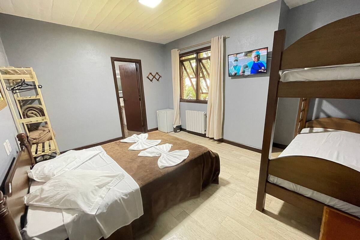 Una habitación con literas, una escalera de madera, una mesita, una silla, un televisor en la pared y una ventana con cortinas.