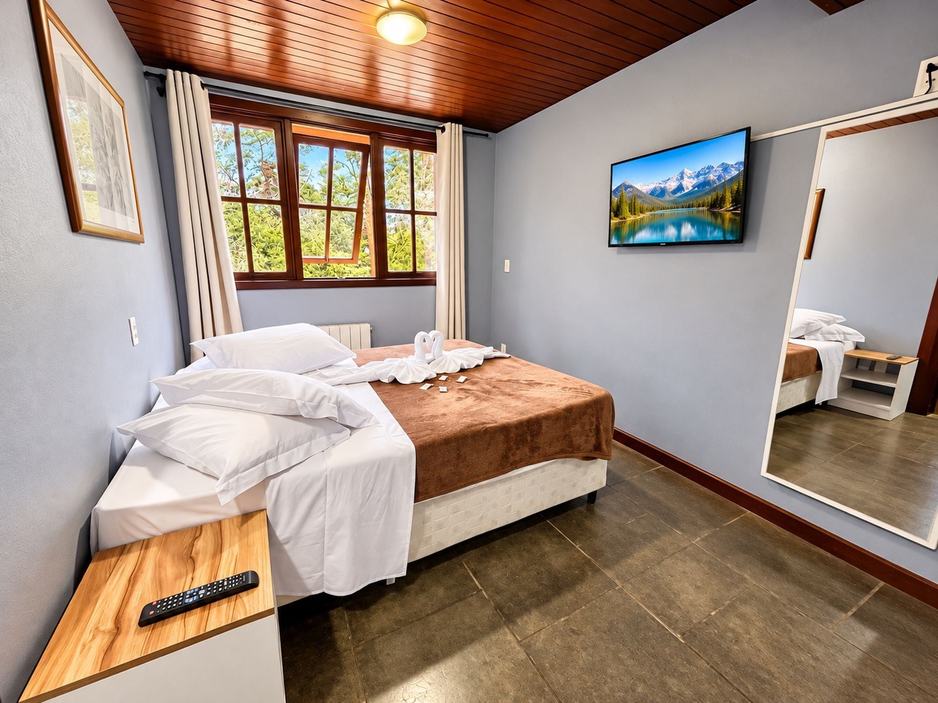 Een slaapkamer met een bed, een nachtkastje met een afstandsbediening, een raam met gordijnen, een schilderij aan de muur en een spiegel die een deel van de kamer weerkaatst.
