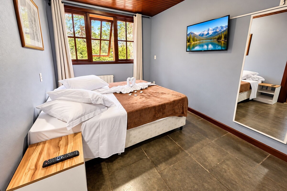 A bedroom with a bed, a bedside table with a remote control, a window with curtains, a painting on the wall, and a mirror reflecting part of the room.