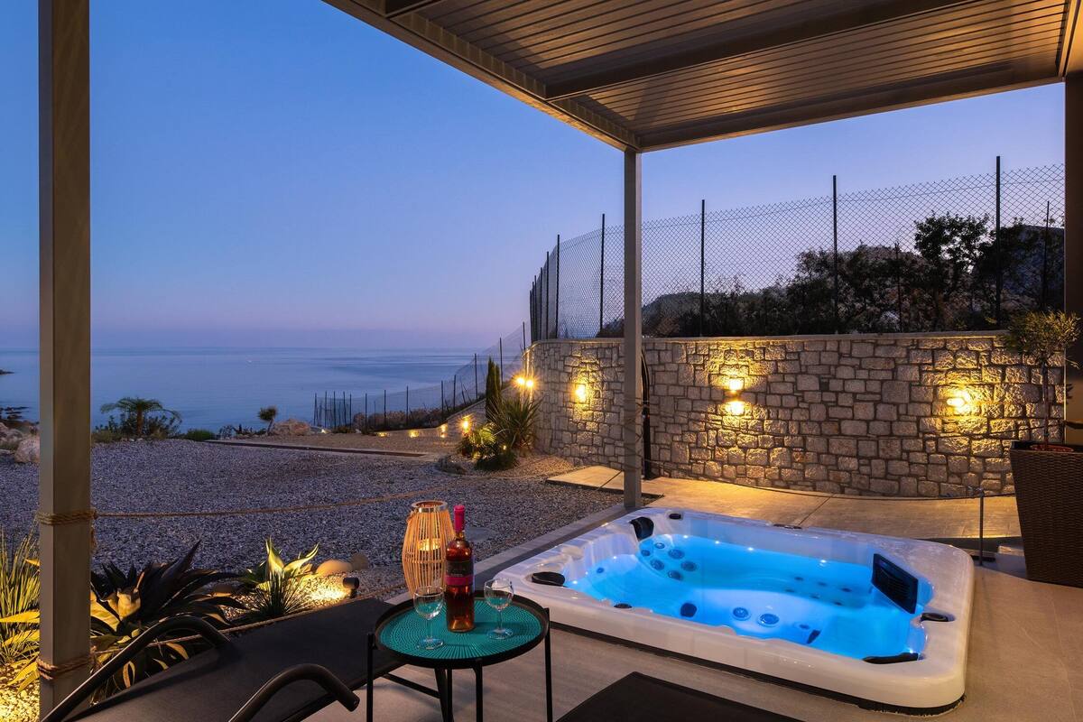 A hot tub with a view of the sea, a table with a bottle and glasses, and a stone wall with lights.