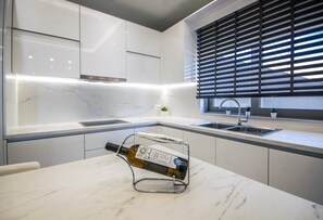 A modern kitchen with white cabinetry, a marble countertop, a wine bottle on a stand, and a window with blinds. - Villa Aquamarine beachfront Rhodes (Stegna)