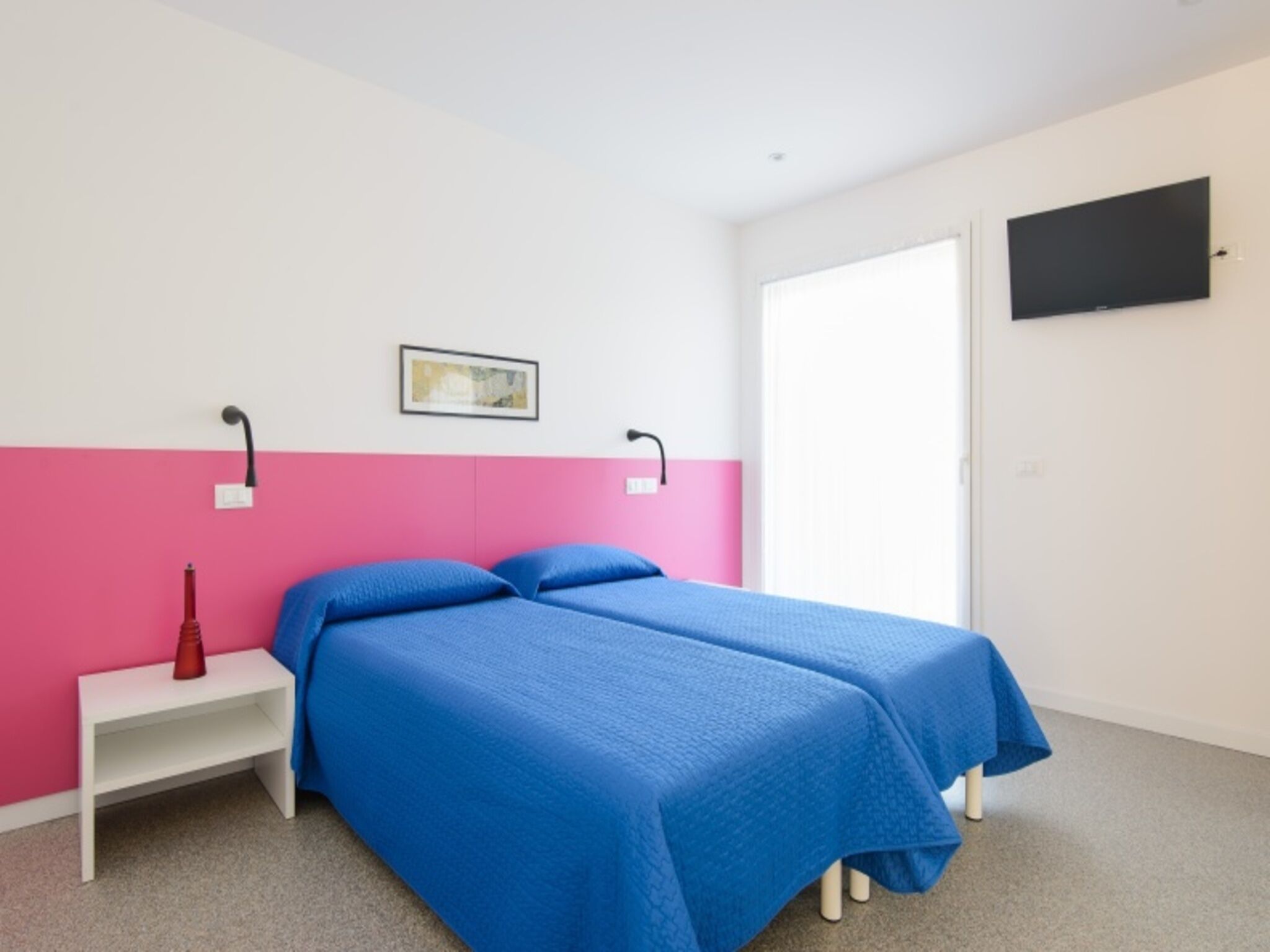 A bedroom with two beds, a white nightstand, a red vase, a painting, and a TV on the wall.