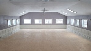 A large, empty room with a high ceiling, multiple windows, and a central ceiling fan.