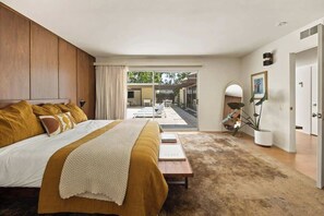 A bedroom with a bed, a bedside table, a lamp, a door, and a view of an outdoor area. - Hugh Kaptur Hideaway Villa (Palm Desert)
