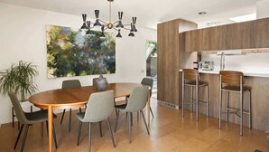 A modern dining area with a round wooden table, grey upholstered chairs, a large abstract painting, and a contemporary chandelier. - Hugh Kaptur Hideaway Villa (Palm Desert)
