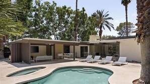 A swimming pool with lounge chairs, a covered patio, and a building with large windows and a flat roof. - Hugh Kaptur Hideaway Villa (Palm Desert)