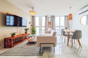 Una sala moderna con un televisor de pantalla plana, un sofá, una mesa de comedor con sillas, una mesa de centro y un ventanal con cortinas.