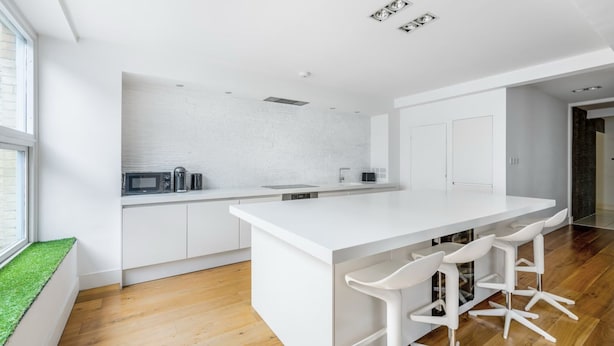 A modern kitchen with white cabinetry, a central island with bar stools, and a window with a green view. - Spacious Modern Apartment in Central London (London)