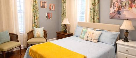 A bedroom with a bed, two chairs, a nightstand, a lamp, and a wall with pictures.