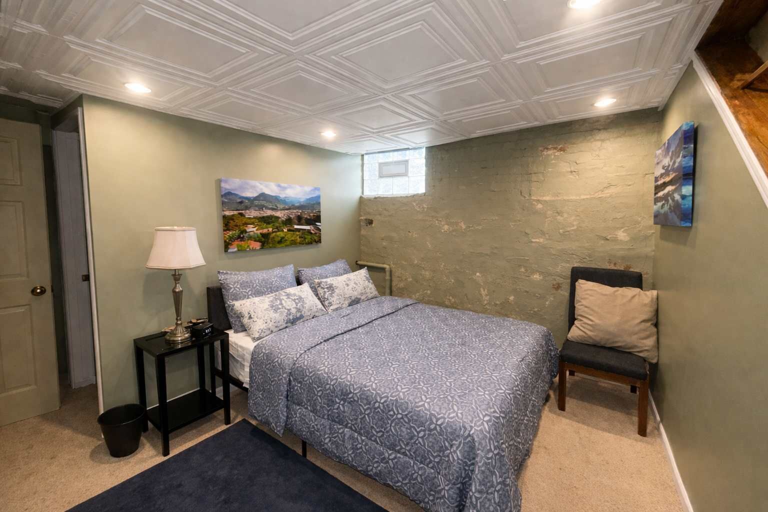 A bedroom with a bed, a nightstand, a lamp, a chair, and a painting on the wall.