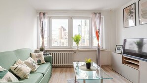 En moderne stue med en grøn sofa, et glasbord, en potteplante, et fjernsyn og udsigt over byen gennem store vinduer.