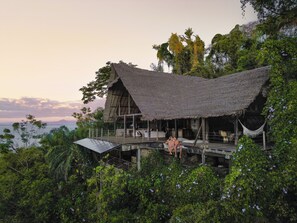 Une construction à toit de chaume, dotée d’une terrasse en bois, entourée d’une végétation luxuriante et offrant une vue sur une étendue d’eau.