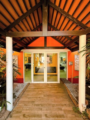 Una entrada techada con piso de baldosas que conduce a un puerta de vidrio, rodeada de plantas y una pared roja.