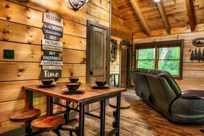 A rustic cabin interior with a wooden wall sign, a dining table with bowls, a leather chair, and a window with a forest view. - Creekside Cottage! Hot Tub, Fire Pit, Pet Friendly (Pigeon Forge)