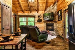 A log cabin interior with a stone fireplace, leather sofa, and a television mounted above it. - Creekside Cottage! Hot Tub, Fire Pit, Pet Friendly (Pigeon Forge)