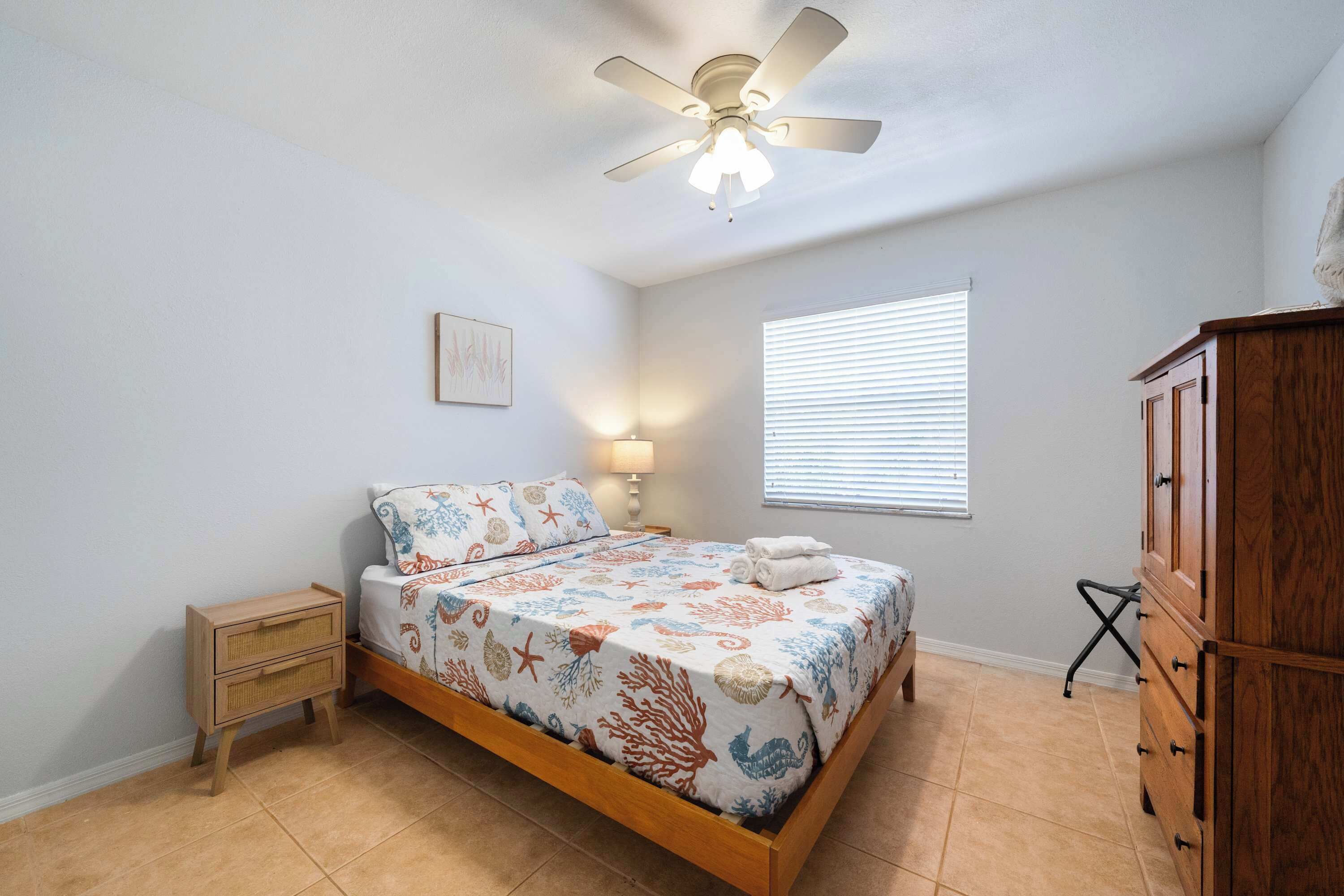 A bedroom with a bed, a nightstand, a dresser, a ceiling fan, a window with blinds, and a wall-mounted lamp.