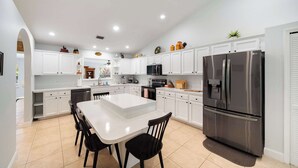 A modern kitchen with white cabinets, a stainless steel refrigerator, and a central island with chairs. - Spacious 5BR Pool Oasis Near Disney Resort (Sebastian)