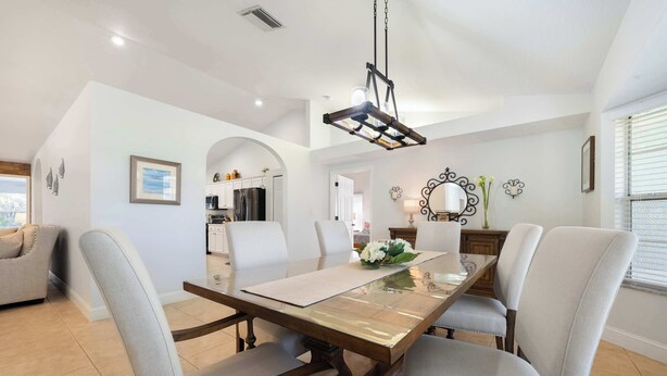 A dining area with a wooden table, cushioned chairs, a chandelier, and a kitchen in the background. - Spacious 5BR Pool Oasis Near Disney Resort (Sebastian)