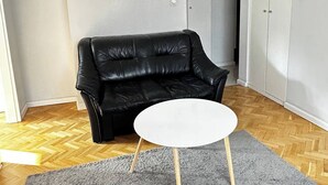Una habitación con un sofá de cuero negro, una mesa redonda blanca con patas de madera y una alfombra gris sobre piso de madera.