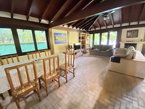 A dining area with wooden chairs and a table, a living room with a sofa and a fireplace, and large windows with a view of greenery. - Casa Jataí (São Sebastião)