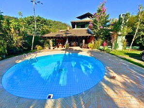 Uma piscina com um deck ladrilhado, rodeada por vegetação exuberante e um edifício tradicional com telhado em azulejos.