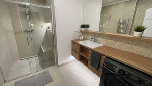 Un bany modern amb plat de dutxa de vidre, un moble de bany de fusta amb rentamiques, un mirall i una màquina de rentar.