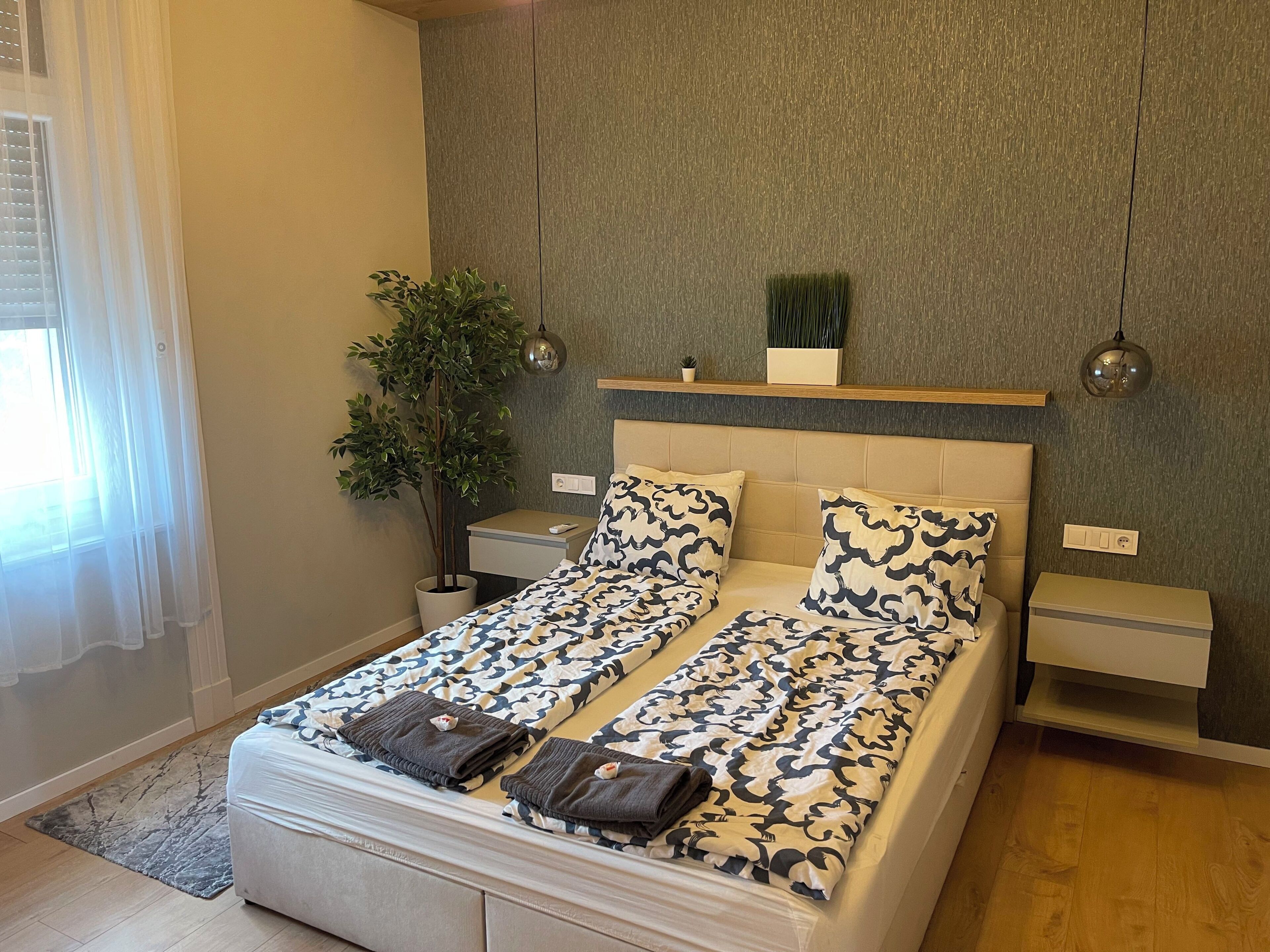 A modern bedroom with a bed, bedside tables, a nightstand, a potted plant, and a window with curtains.