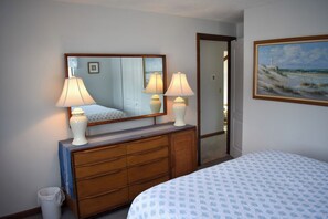Quarto com cama, penteadeira, espelho, duas luminárias, um quadro e uma porta que dá para outro cômodo.