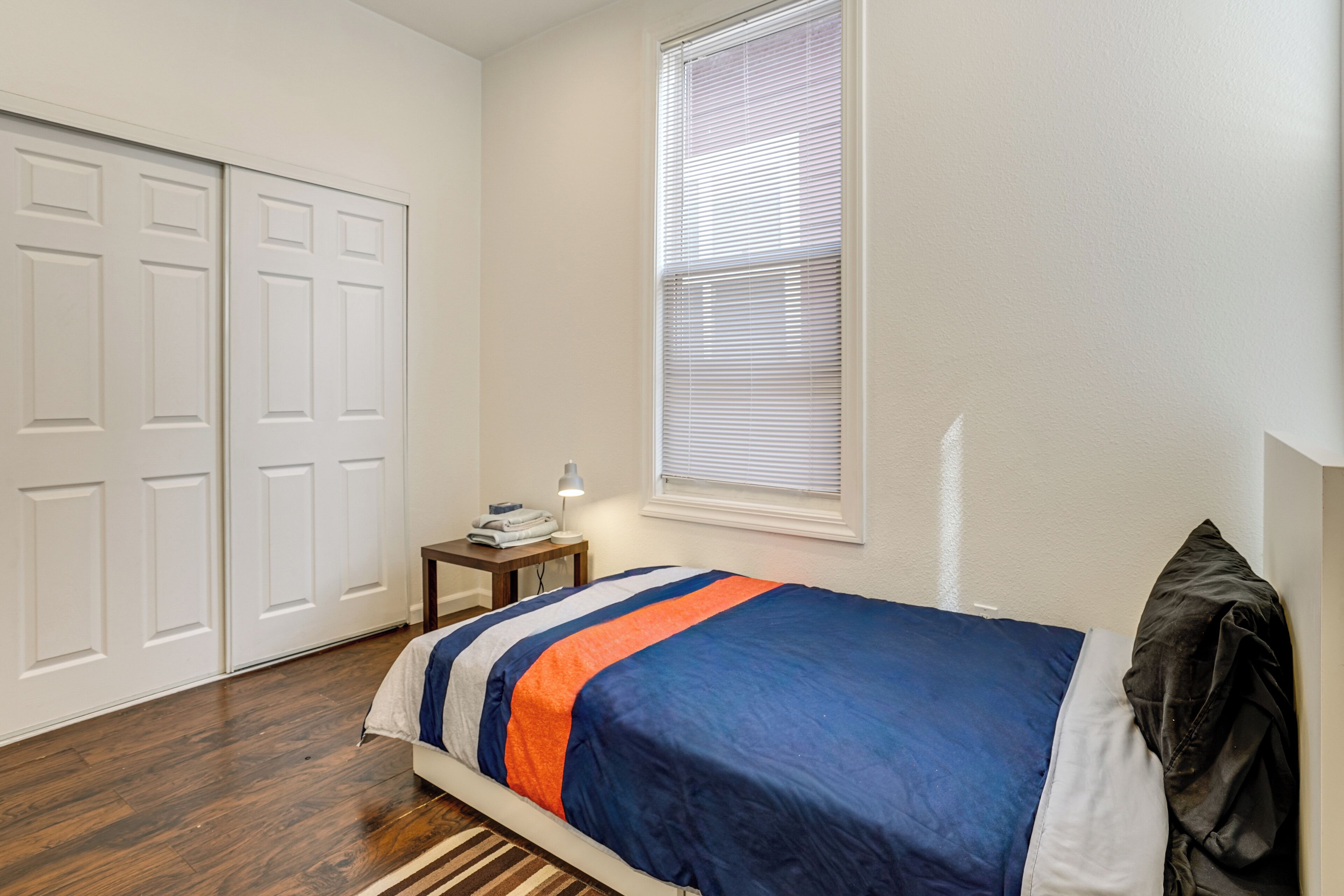 Un dormitorio con cama, escritorio, lámpara, ventana con persianas y armario.