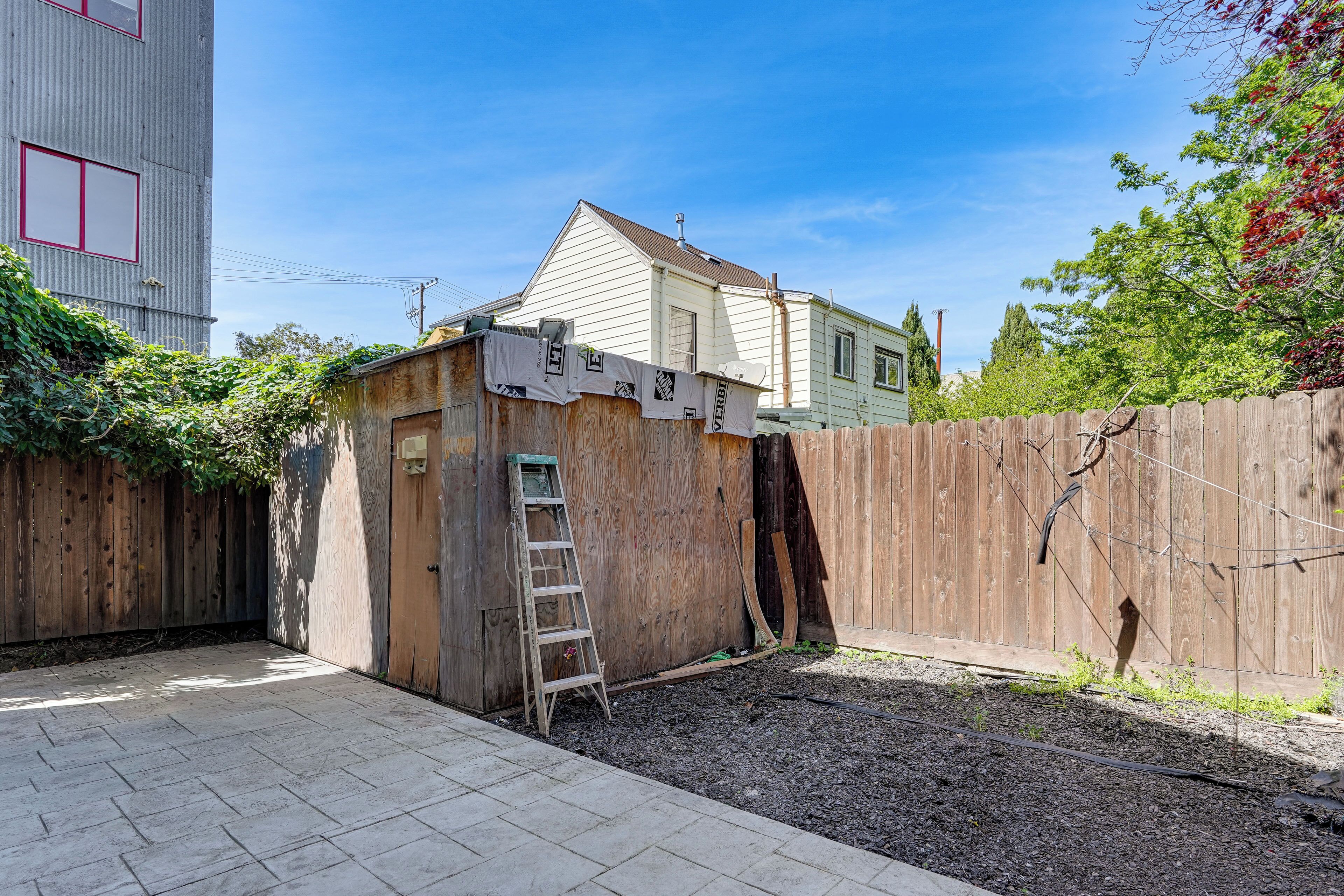 A fenced backyard with a wooden shed, a ladder, and a house in the background.