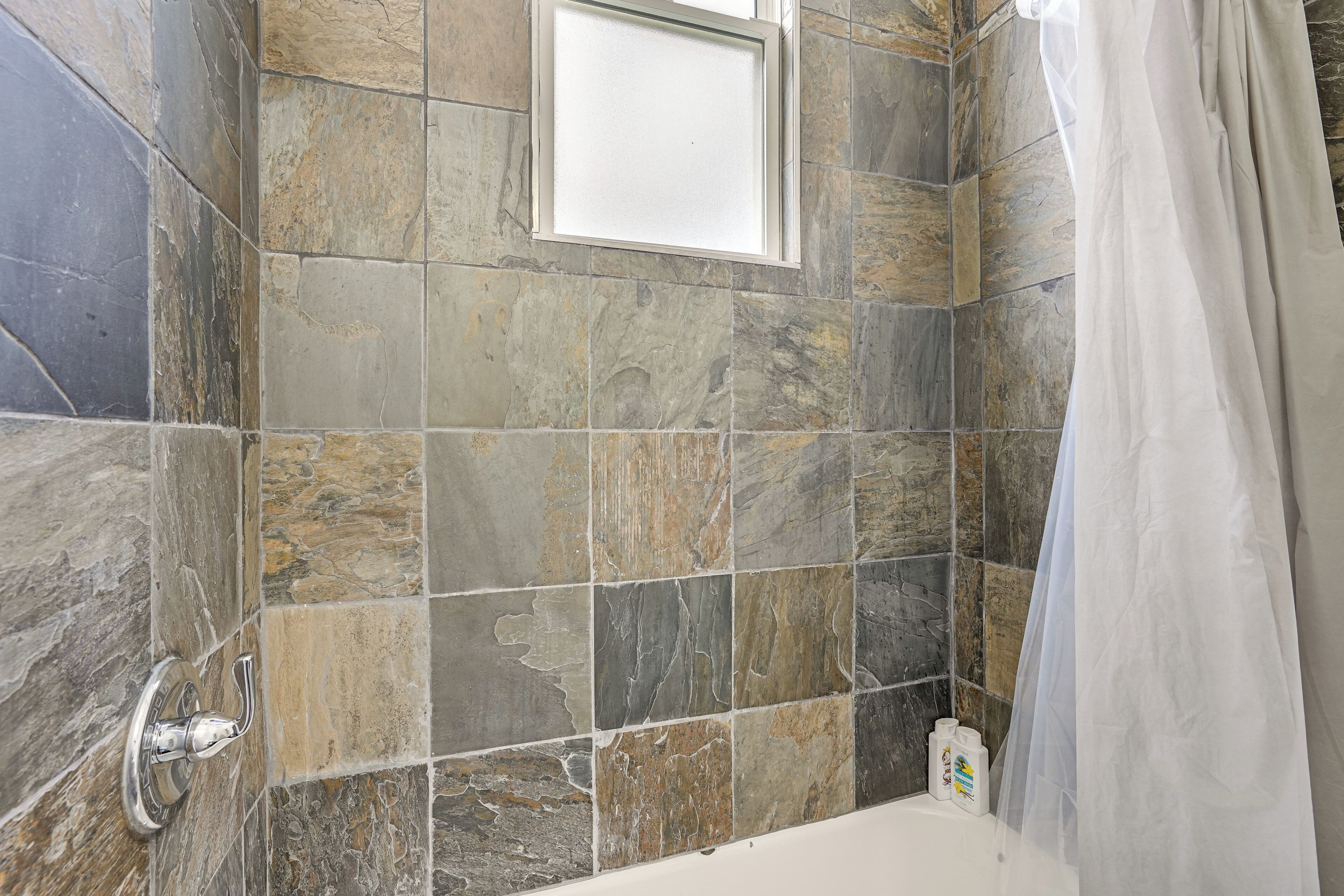 Un bagno con piastrelle in pietra, una finestra, una tenda da doccia e una bottiglia di shampoo.