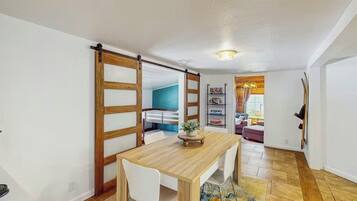 Un coin repas avec une table et des chaises en bois, une porte coulissante et une vue sur une autre pièce dont le mur est bleu et qui contient une bibliothèque.