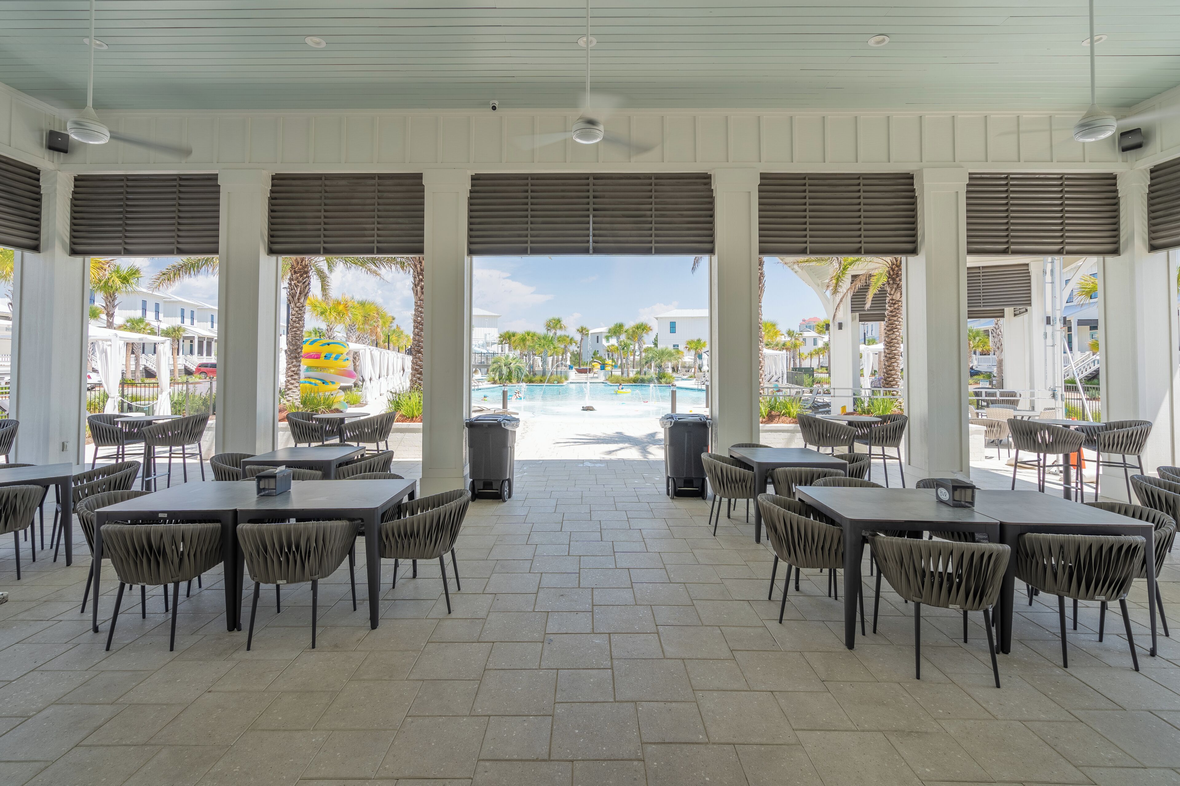 An outdoor dining area with tables and chairs, a view of a pool, and palm trees.