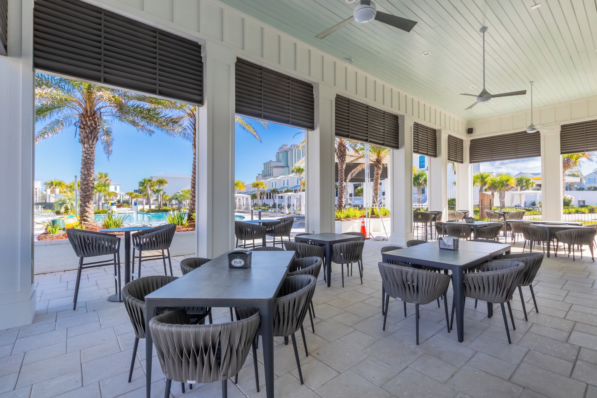 An outdoor patio with tables and chairs, large windows with blinds, and a view of a pool and buildings.