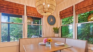 A dining area with a wooden table and bench, large windows with bamboo blinds, a hanging light fixture, and a wall clock. - Peaceful Mountain Hideaway with a view, near Maggie Valley and Asheville (Waynesville)