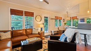 A living room with a brown leather sofa, a black armchair, a coffee table, and a patterned rug. There is a ceiling fan, a dining area with a table and chairs, and a kitchen area with a countertop and a hanging light fixture. - Peaceful Mountain Hideaway with a view, near Maggie Valley and Asheville (Waynesville)