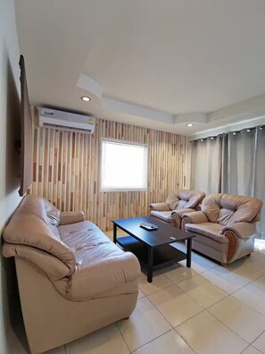 Ein Wohnzimmer mit zwei beigen Sofas, einem Couchtisch, einem an der Wand montierten Fernseher und einem Fenster mit Jalousien.