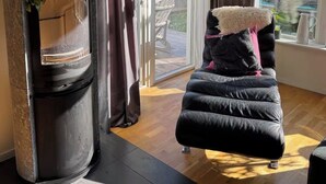 Ein Zimmer mit Kamin, gepolstertem Sessel, einer Topfpflanze und großen Fenstern.