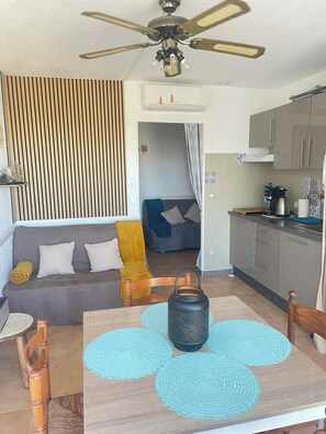 Een moderne woonkamer met een bank, een eettafel met placemats, een plafondventilator en een keuken met kastjes en een koffiezetapparaat.