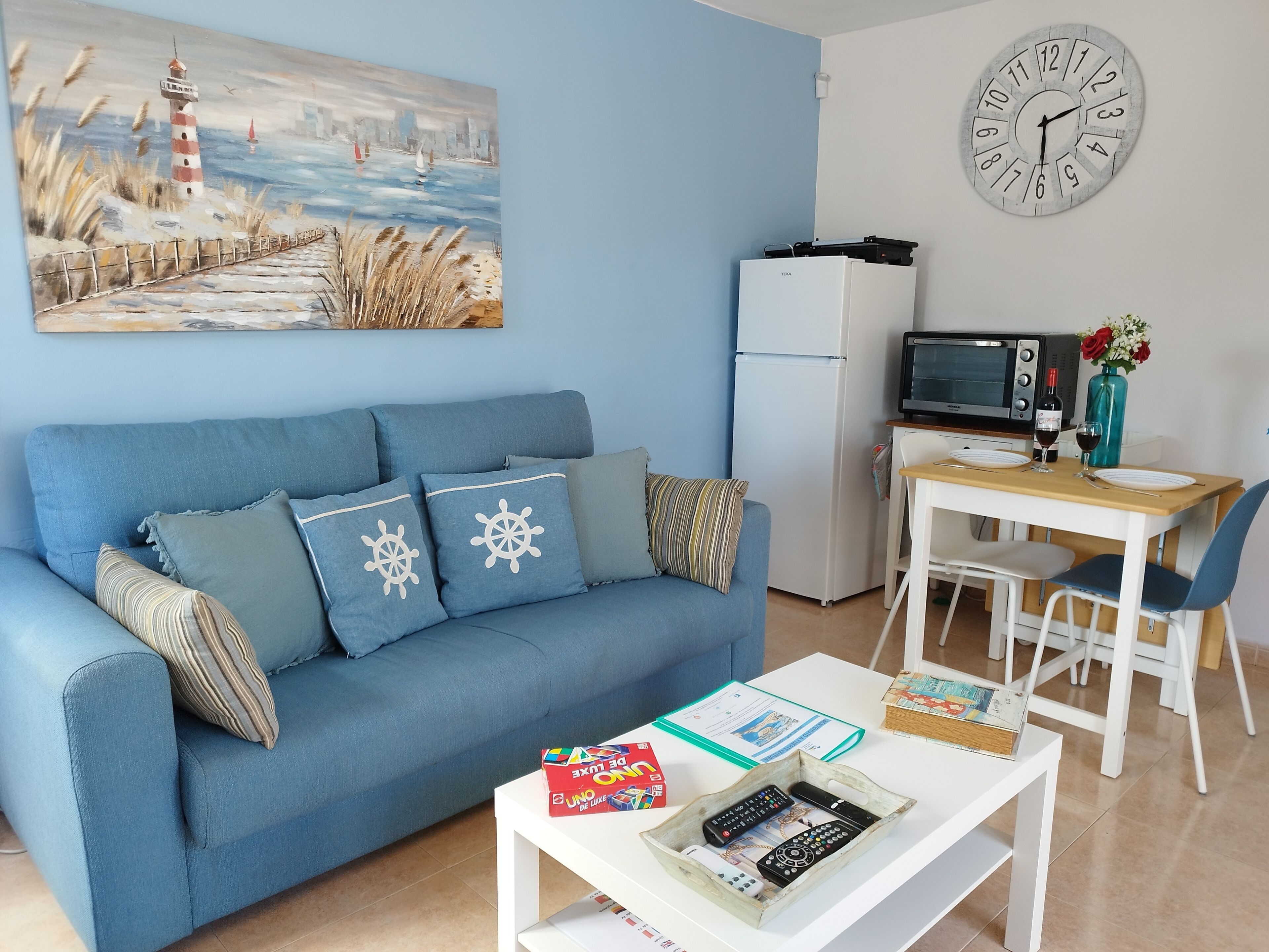 Ein Wohnzimmer mit blauem Sofa, einem Gemälde eines Leuchtturms, einer weißen Uhr, einem Esstisch mit Stühlen, einem Fernseher und einem Kühlschrank.