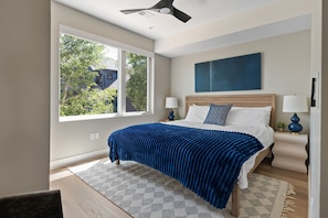 A bedroom with a large window, a bed with a blue blanket, two bedside lamps, and a blue abstract painting on the wall. - Moose Hollow 3 Bedroom Home (Winter Park)