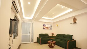 A modern living room with a sectional sofa, a small wooden table, recessed ceiling lighting, and a decorative shelf. - Krishna Royal Club (Guruvayur)